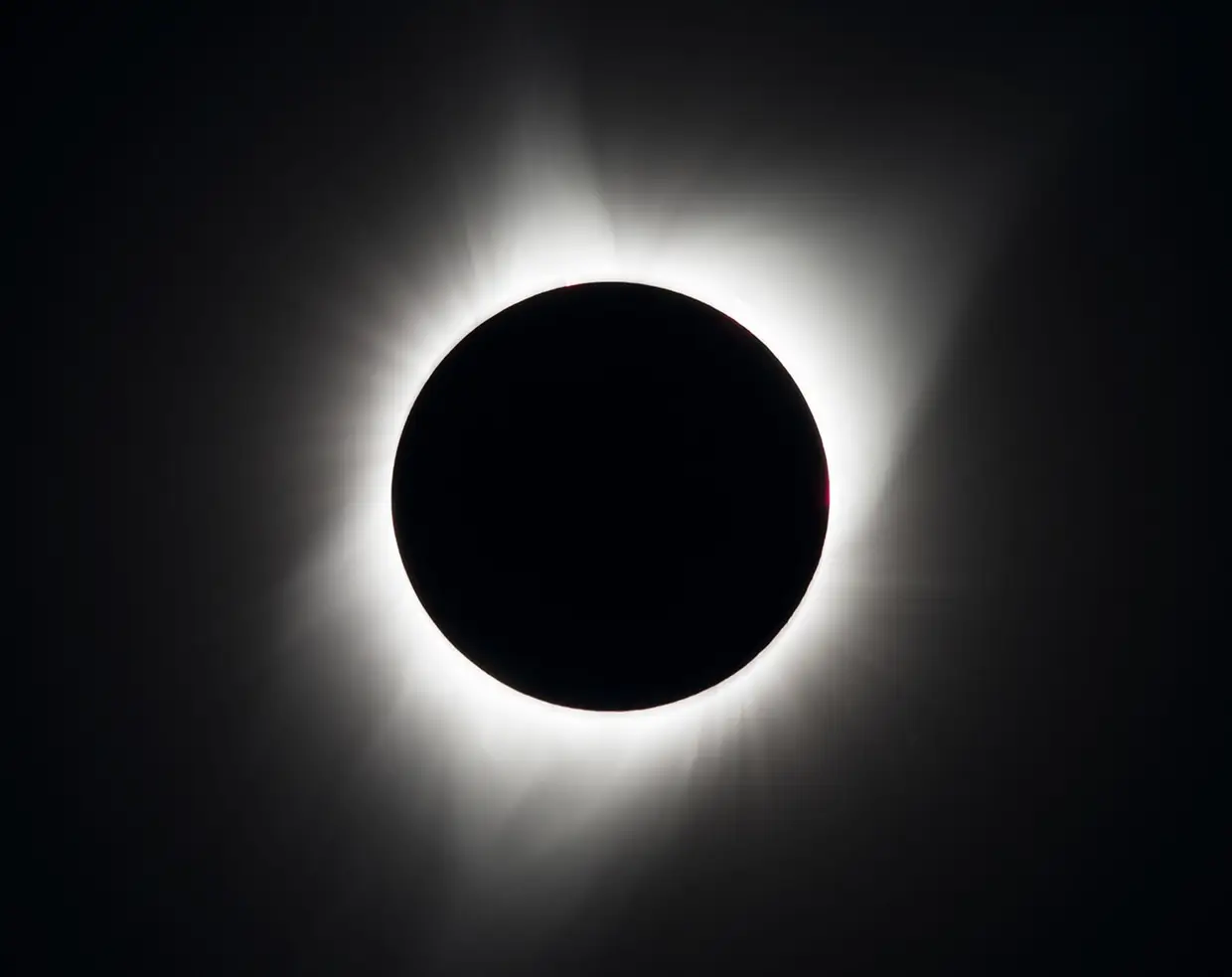 The Sun's white corona streaming outwards around the black disc of the Moon, with two small pink prominences at the edge, against a dark sky.