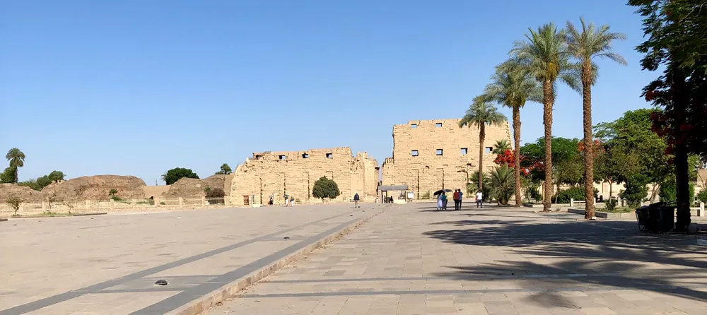 The first pylon of Karnak Temple across a wide paved esplanade, with date palms to the right and a handful of visitors.